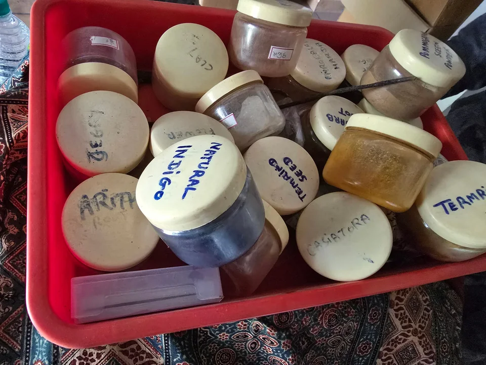 A tray of jars of natural dye stuff, lids marked by hand: natural indigo, tamarind seed, cassia tora, clay, pomegranate seed powder, on ajrakh cloth