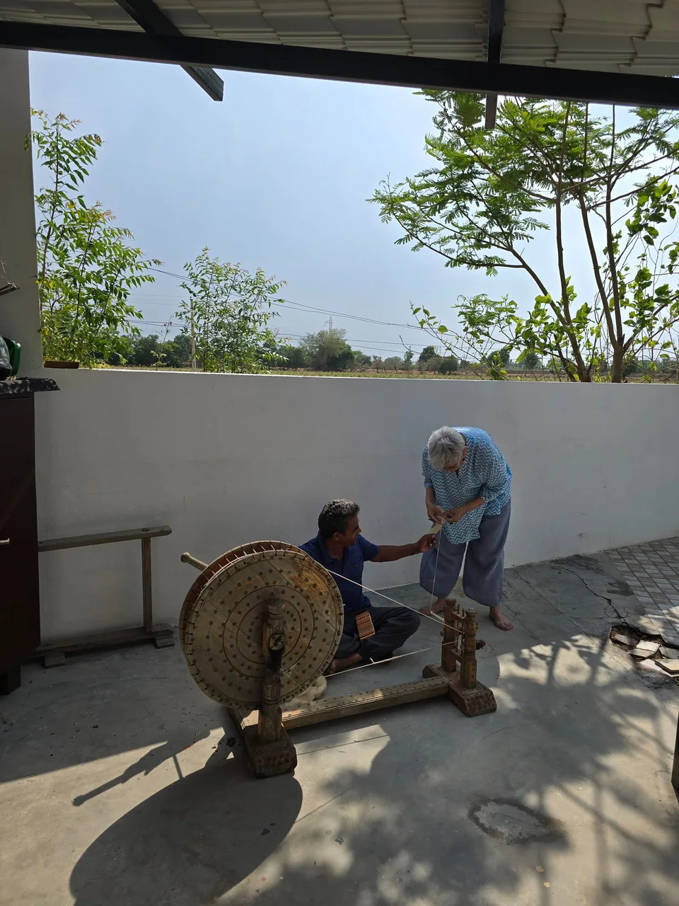 Two artisans at a large wooden wheel in a shaded courtyard, trees beyond