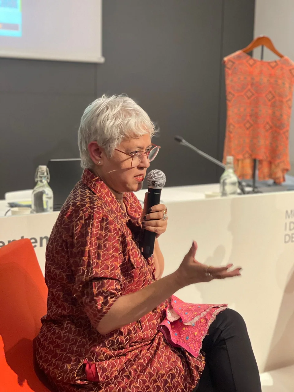 Manjula Sen speaking with a microphone at the Museu Etnologic i de Cultures del Mon in Barcelona, a red Sensclo garment on a stand behind her
