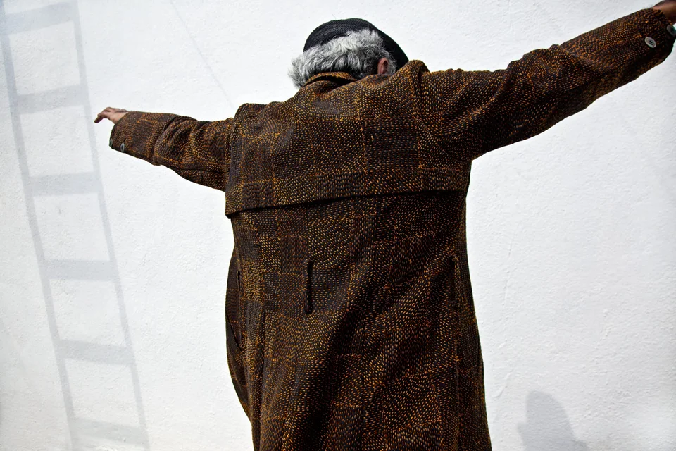 Mahesh Balasubramaniam in a long brown coat, seen from behind with arms stretched out