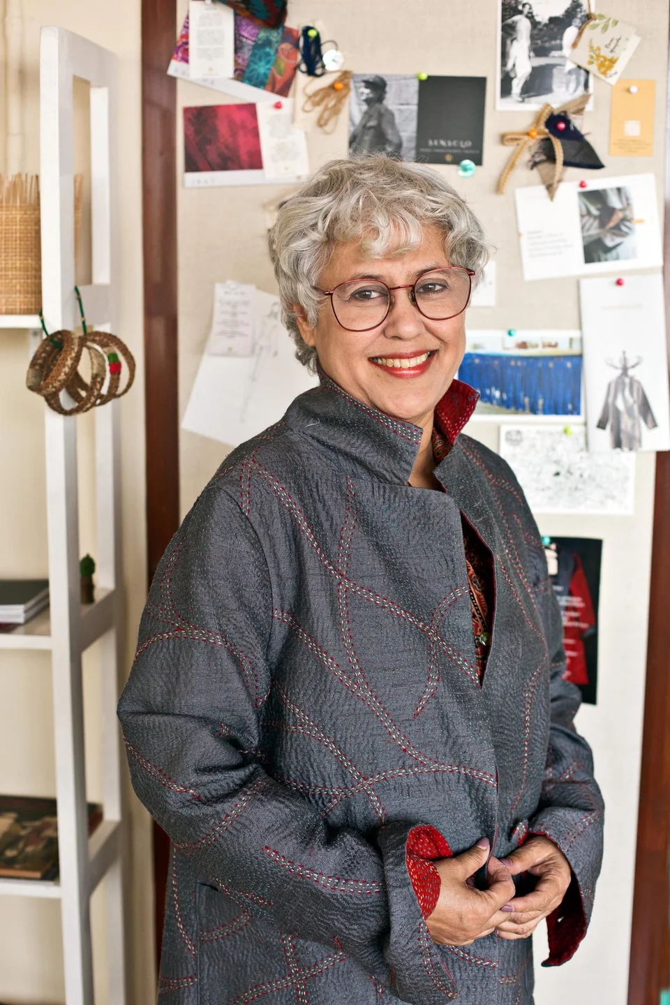 Manjula Sen in her studio in a grey kantha coat, standing in front of her working wall of sketches and swatches